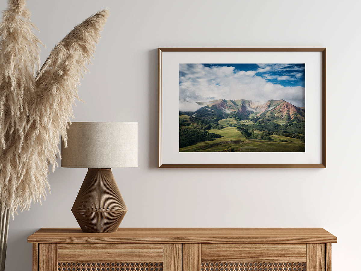 Stunning landscape photograph of the green, rolling hills and majestic mountains of Crested Butte under a bright blue sky with scattered white clouds.