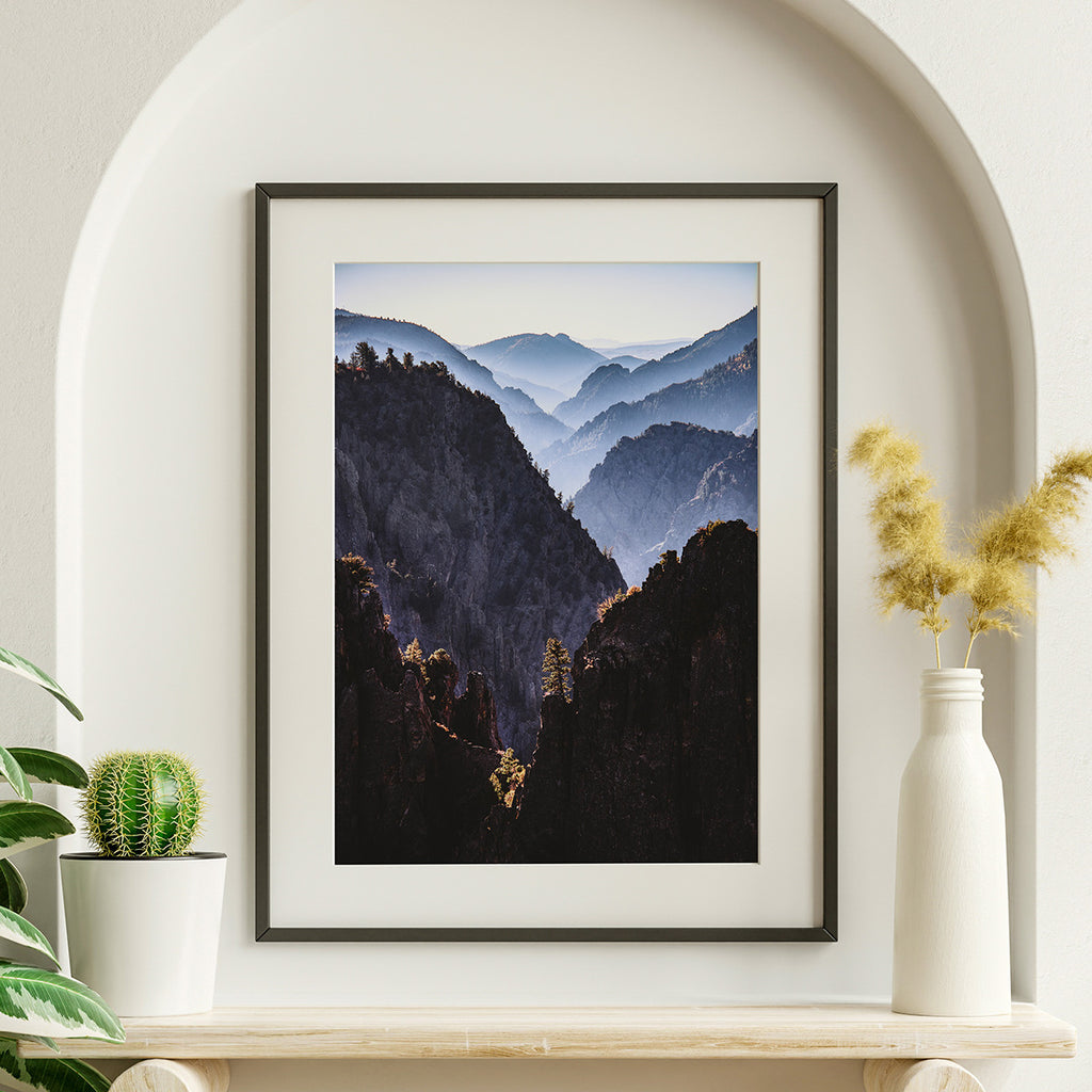 Landscape photograph of a deep canyon with layered mountains fading into the distance, warm sunlight on foreground rocks, lone tree in center, muted blues and grays.