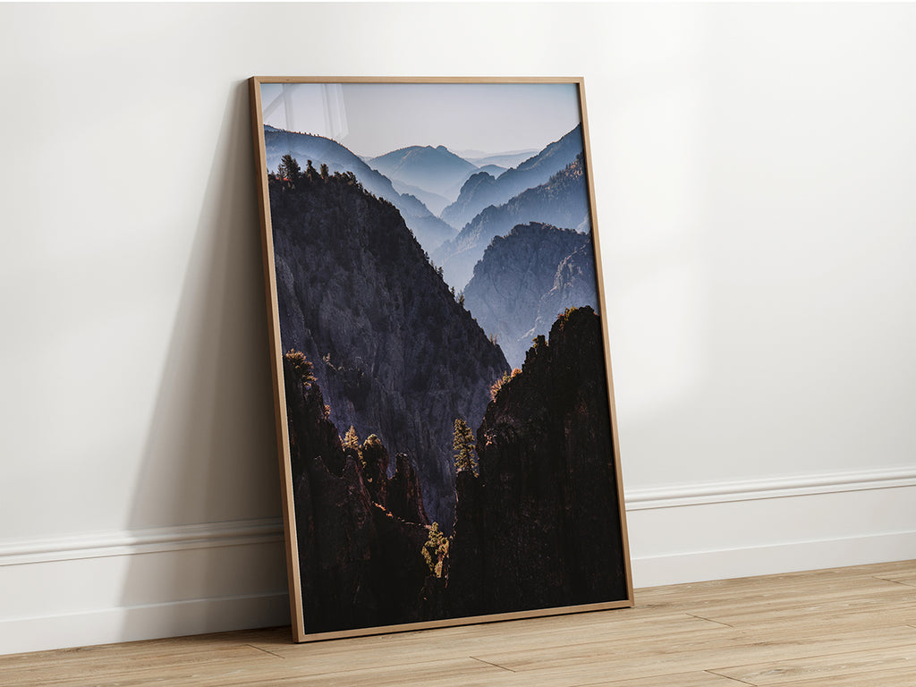 Landscape photograph of a deep canyon with layered mountains fading into the distance, warm sunlight on foreground rocks, lone tree in center, muted blues and grays.