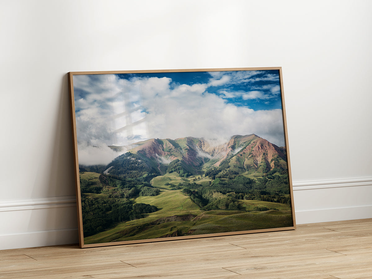 Stunning landscape photograph of the green, rolling hills and majestic mountains of Crested Butte under a bright blue sky with scattered white clouds.