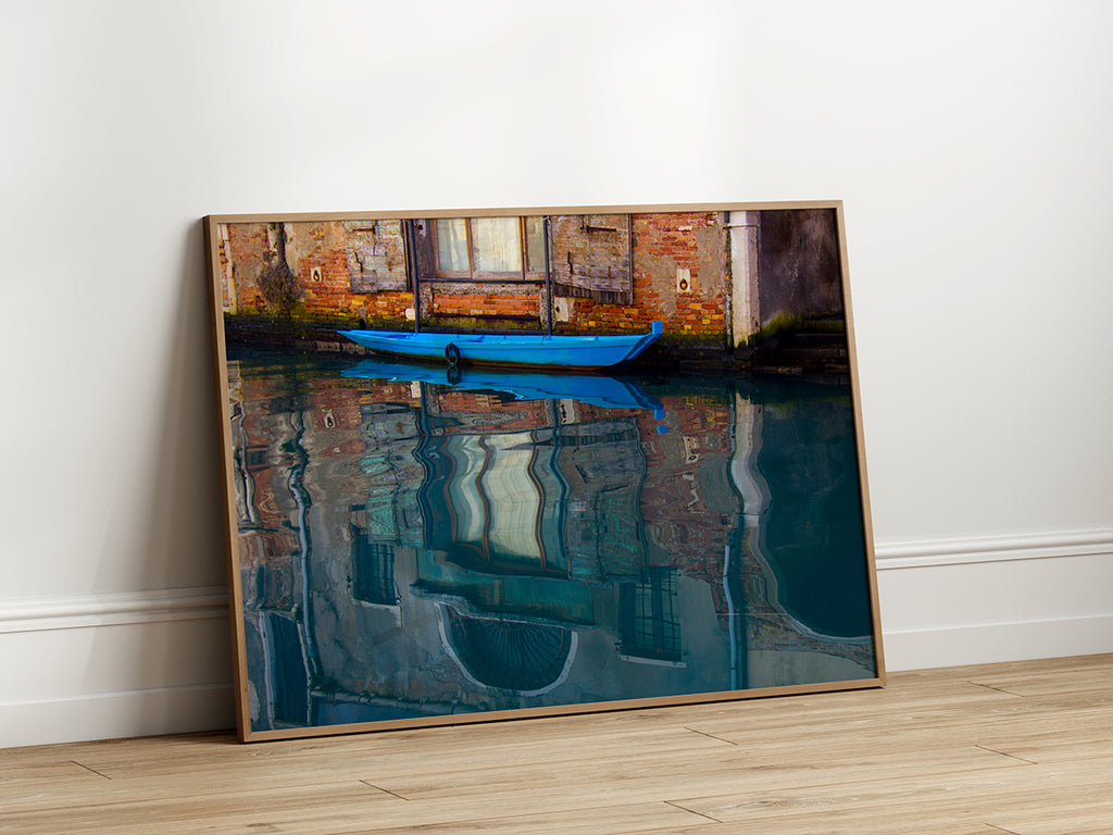 Artwork featuring a bright blue boat tied to the side of a canal in Italy, with detailed reflections of the surrounding brick and stone buildings in the rippling water.