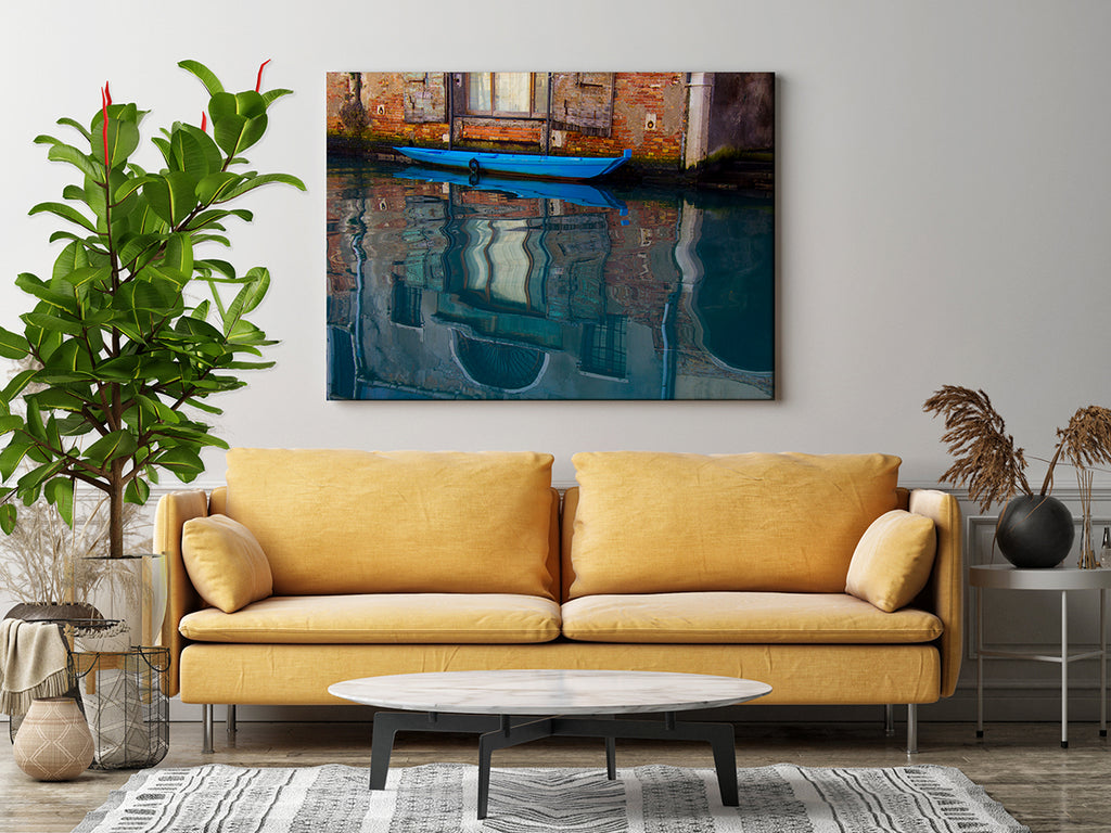 Artwork featuring a bright blue boat tied to the side of a canal in Italy, with detailed reflections of the surrounding brick and stone buildings in the rippling water.